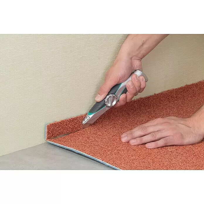 Wolfcraft Trapezmesser - Länge: 52 mm - Eine Person schneidet orangefarbenen Teppich mit einem Teppichmesser bei der Verlegung in einer Raumecke.