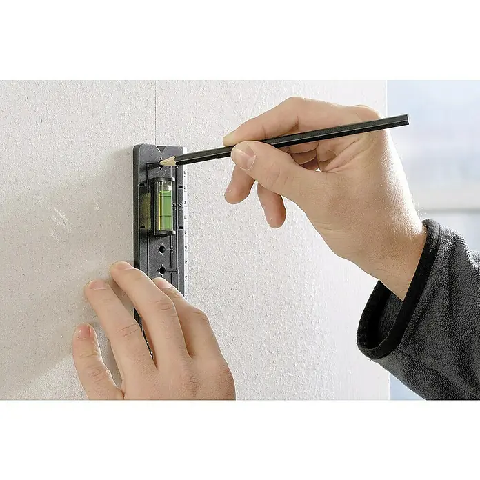 Wolfcraft Plantilla de perforación - 185 x 50 mm, Apto para: Cajas para empotrar - Un hombre marca un agujero en una pared con un lápiz, utilizando un nivel de burbuja para la alineación. Precisión en bricolaje.
