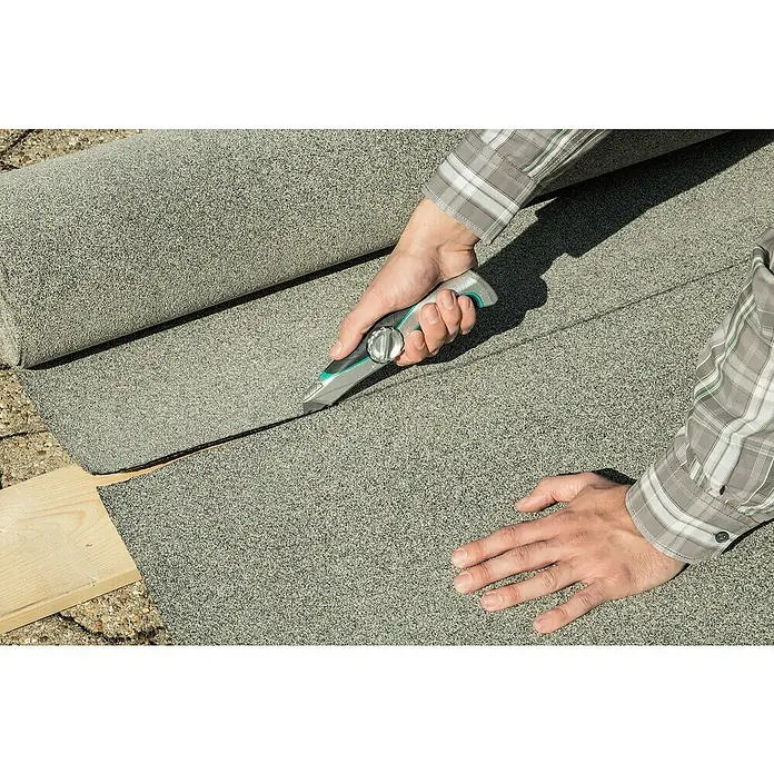 Wolfcraft Trapezmesser - Länge: 52 mm - Ein Mann schneidet Dachpappe mit einem Cuttermesser auf einer Holzunterlage. Das Material ist grau und gerollt.