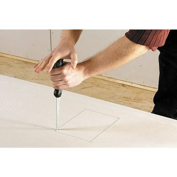 Wolfcraft Serrucho de calar para placas de yeso laminado - Apto para: Placas de yeso laminado - Un hombre usa una sierra de calar para cortar un agujero cuadrado en paneles de yeso, resaltando la herramienta y el material trabajado.