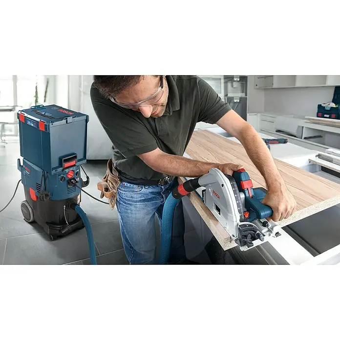 Bosch Professional Tauchsäge GKT 55 GCE - 1.400 W, Durchmesser Sägeblatt: 165 mm, Leerlaufdrehzahl: 6.250 U/min - Ein Handwerker sägt mit einer Bosch Tauchsäge ein Holzbrett, angeschlossen an einen Bosch Industriesauger für eine saubere Arbeitsumgebung.