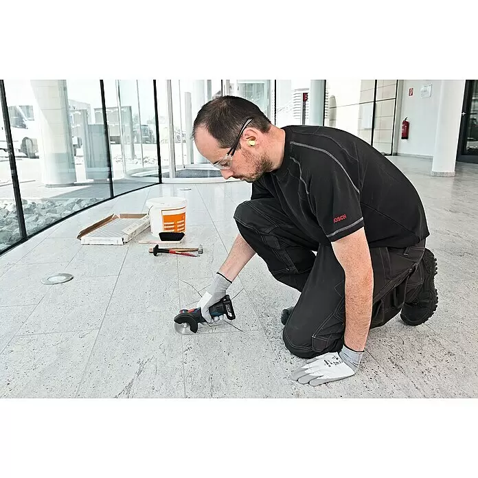 Bosch Professional Akku-Winkelschleifer GWS 12V-76 - 18 V, 2 Akkus, 2 Ah, Durchmesser Scheibe: 76 mm - Mann in Schutzbrille und Gehörschutz verwendet einen blauen und schwarzen Akku-Winkelschleifer zum Schneiden von Bodenfliesen.