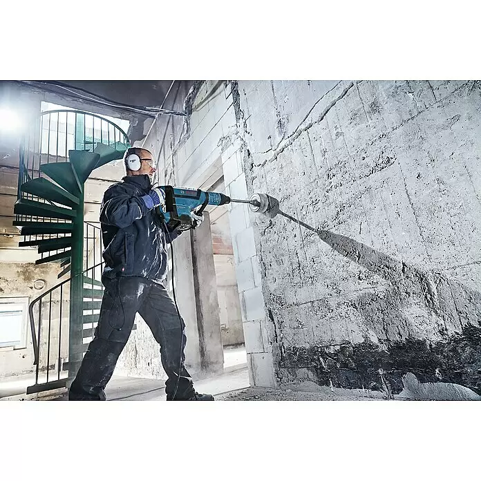 Bosch Professional Kombihammer GBH 12-52DV - 1.700 W, 19 J, SDS-Max-Aufnahme - Eine Person bohrt mit einem blauen Bosch Bohrhammer, Gehörschutz und Schutzbrille ein großes Loch in eine Betonwand auf einer Baustelle.
