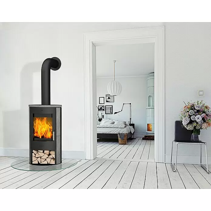 Fireplace Kaminofen Zaria - 6,6 kW, Raumheizvermögen: 108 m³, Material Verkleidung: Speckstein, Schwarz - Moderner grau-schwarzer Kaminofen mit sichtbaren Flammen und Holzscheiten, auf Glasplatte in Wohnraum.