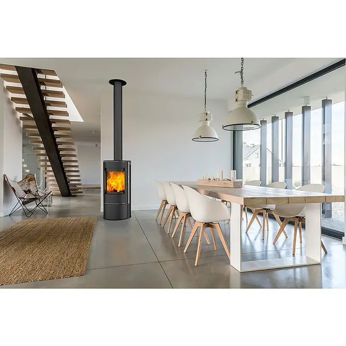 Fireplace Kaminofen Rondale - 5,5 kW, Raumheizvermögen: 90 m³, Material Verkleidung: Stahl, Schwarz - Das Bild zeigt einen modernen Kaminofen mit loderndem Feuer, der Wärme und Atmosphäre in den Raum bringt. Elegante Einrichtung mit Esstisch und Stühlen.