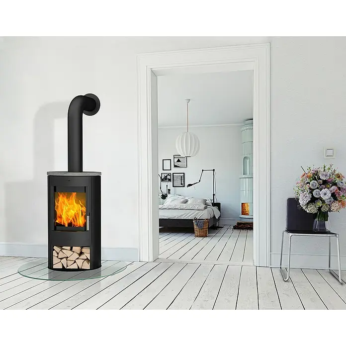 Fireplace Kaminofen Zaria - 6,6 kW, Raumheizvermögen: 108 m³, Material Verkleidung: Speckstein, Schwarz - Ein moderner Kaminofen in Schwarz mit Holzscheiten, der Wärme und Stil in ein helles, minimalistisches Interieur bringt.
