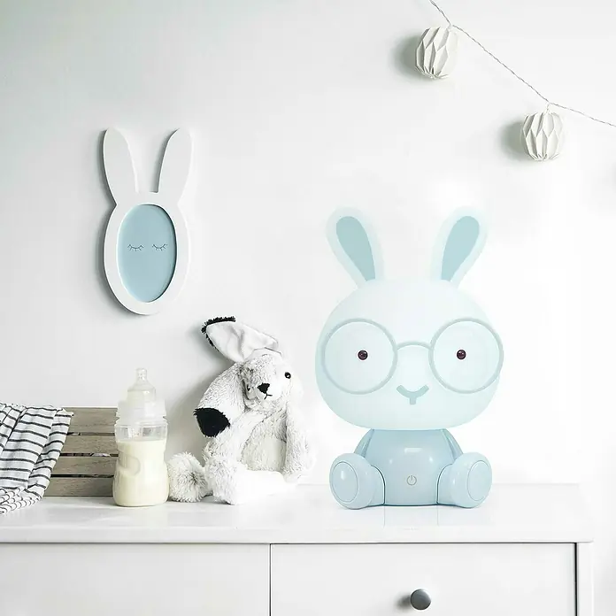 Idp Lampshades Lámpara de sobremesa LED Bunny - 3 W, Ø x Al: 19,5 x 30 cm, Azul - Escena de habitación infantil con una lámpara de conejo con gafas, un peluche, un biberón y un marco con orejas de conejo.