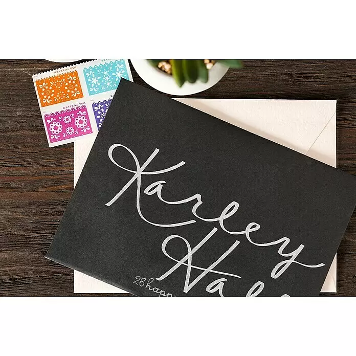Cricut Joy Gelroller-Set Medium Gel Pen Glitter - 3 Stk., Glitzerschwarz/Glitzergold/Glitzersilber, 0,8 mm - Schwarzer Umschlag mit weißer Kalligrafie, teils über cremefarbenem Umschlag, neben bunten Briefmarken.