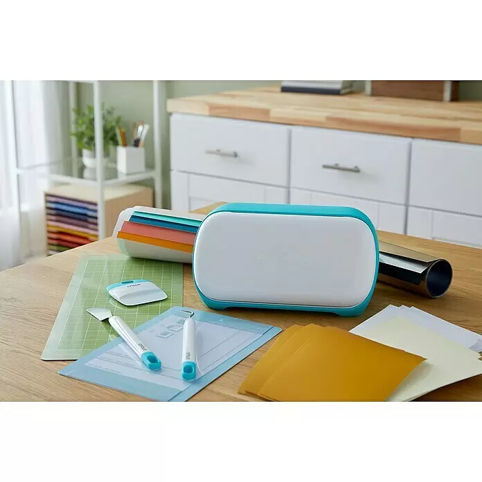 Cricut Joy Plotter-Werkzeug Starter-Werkzeugset - 3 -tlg., Passend für: Cricut Joy Schneideplotter - Eine weiße und türkisfarbene Cricut Joy Maschine auf einem Holztisch, umgeben von Bastelmatten, Werkzeugen und Materialien, bereit zum Basteln.