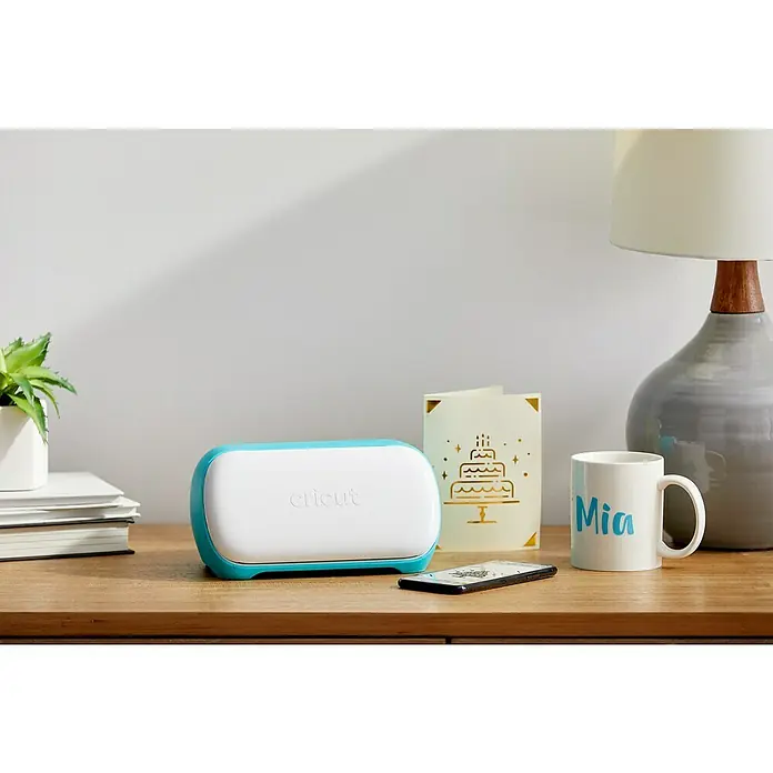 Cricut Joy Schneideplotter Smarte Schneide- und Zeichenmaschine - Türkis/Weiß, Bluetooth, Schnittbreite: 10,4 cm, Schnittlänge: 1,2 m - Nahaufnahme eines Cricut Joy Schneideplotters auf einem Holztisch neben Dekoartikeln.