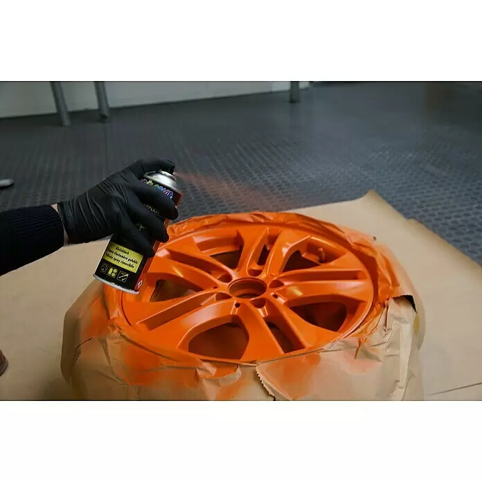 Dupli-Color Tuning Abziehlack SPRAYPLAST - Orange, 400 ml - Behandschuhte Hand sprüht eine orangefarbene Autofelge mit einer bunten Sprühdose, teilweise abgedeckt.