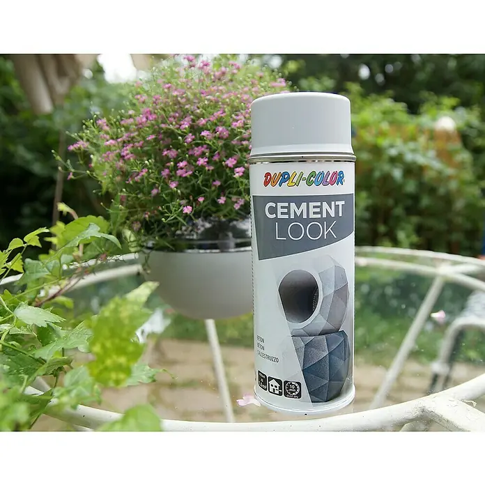 Dupli-Color Spezial-Lackspray Cement Look - Hoover Dark, Matt, 400 ml - Nahaufnahme einer Dupli-Color Zement-Look-Sprühdose auf einem Tisch im Freien, umgeben von Pflanzen und Blumen.