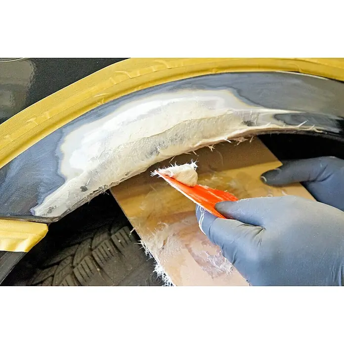 Presto Glasfaserspachtel Prestolith extra - 250 g - Auftragen von Glasfaserspachtel auf einen schwarzen Auto-Radlauf mit einem roten Spachtel, um Schäden zu reparieren.