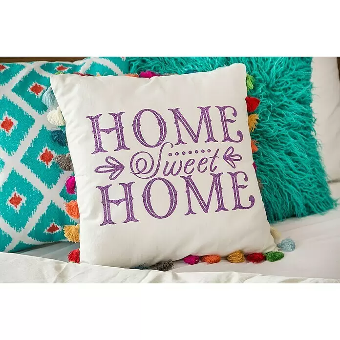 Cricut Kissenhülle Rohling für Infusible Ink - Weiß, L x B: 45,5 x 45,5 cm, 100 % Polyester - Weißes Zierkissen mit lila Glitzerschrift "HOME Sweet HOME" und bunten Quasten, auf einem Bett liegend.
