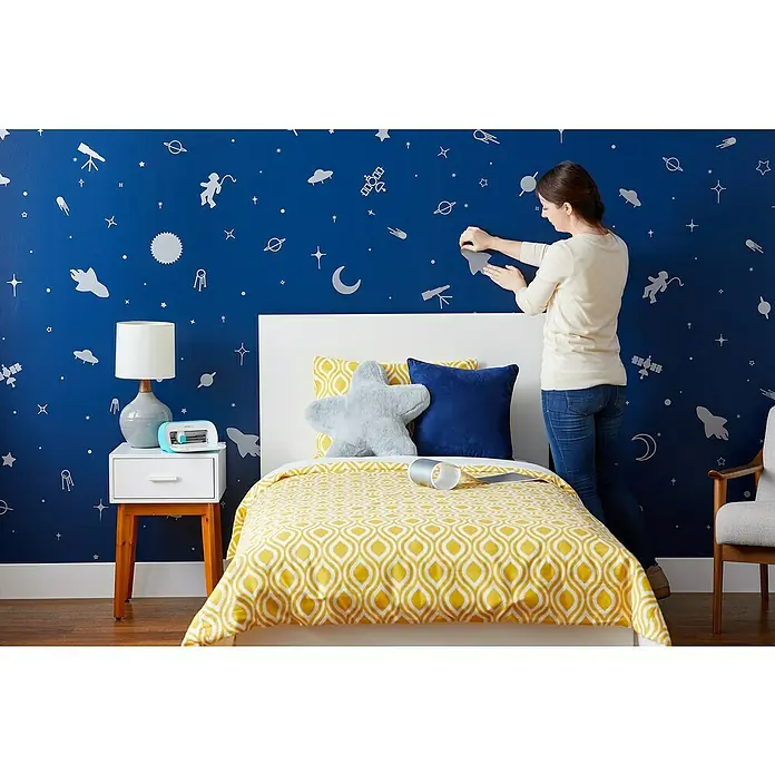 Cricut Joy Vinylfolie Smart Vinyl Ablösbar - Silber, 121,9 x 13,9 cm, Wieder entfernbar - Ein Kinderzimmer mit einer blau bemalten Wand mit weißen Weltraumaufklebern und einem Bett mit gelbem Bettbezug.