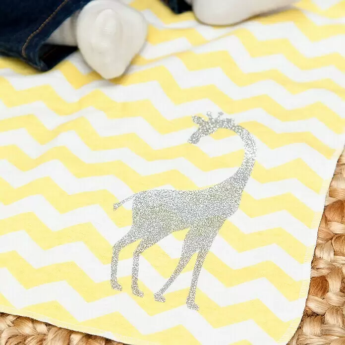 Cricut Aufbügelfolie Glitter Iron-On - 3 Stk., Gold/Silber/Schwarz, L x B: 30,5 x 30,5 cm, Passend für: Cricut Schneideplotter - Babydecke mit gelbem und weißem Chevron-Muster und einer silbernen Glitzer-Giraffe. Beine einer Person ruhen darauf.