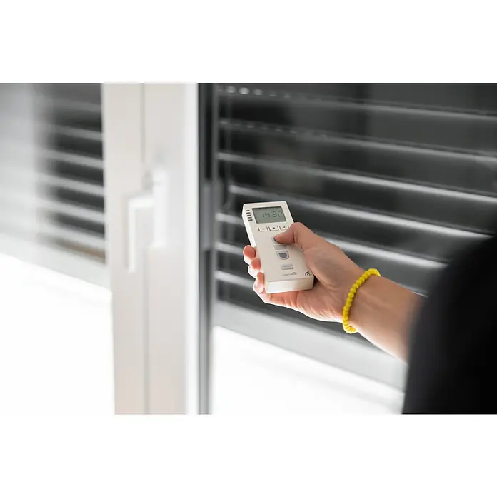 Schellenberg Temporizador inalámbrico Premium - Sistemas soportados: Sistema Smart Home Schellenberg, Apto para: 25 motores telecontrolados - Primer plano de una persona con una pulsera amarilla que sostiene un control remoto blanco para persianas frente a una ventana con persianas.