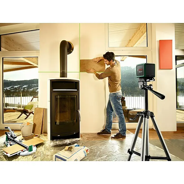 Bosch Kreuzlinienlaser Quigo Green II - Max. Arbeitsbereich: 12 m - Ein Mann verlegt Fliesen mit einem Bosch-Laser-Nivelliergerät an einer Wand neben einem Kaminofen, um präzise und gerade Linien zu gewährleisten.