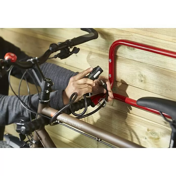 Fahrradhalterung B053QRA - Geeignet für: 2 Fahrräder, Belastbarkeit: 30 kg - Hände sichern ein braunes Fahrrad mit schwarzem Kabelschloss an einem roten Wandhalter an einer Holzwand.