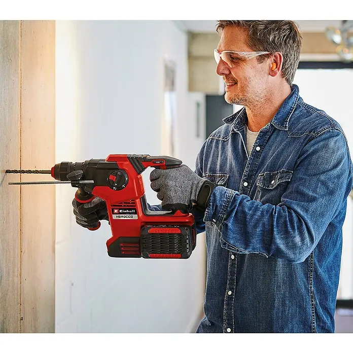 Einhell Professional PXC 18V Martillo combinado de batería Herocco 36/28 - 18 V, Sin batería, 0 Ah, 3,2 J - Un hombre perfora una pared con un taladro percutor inalámbrico Einhell Herocco rojo, usando gafas de seguridad y guantes.