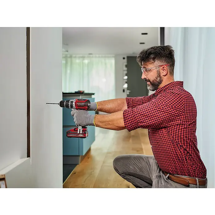 Einhell PXC 18V Taladro atornillador percutor de batería TP-CD 18/60 Li- i BL - 18 V, Sin batería, 60 Nm - Un hombre atornilla un tornillo en una pared con un taladro atornillador Einhell. Seguridad con gafas protectoras y guantes durante el bricolaje.