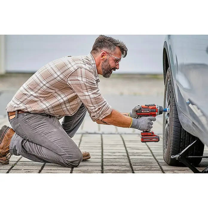 Einhell Professional PXC 18V Llave de impacto de batería IMPAXXO 18/400 - 18 V, Sin batería, 450 Nm - Un hombre se arrodilla utilizando una llave de impacto inalámbrica Einhell roja para trabajar en un coche, con gafas de seguridad y guantes.