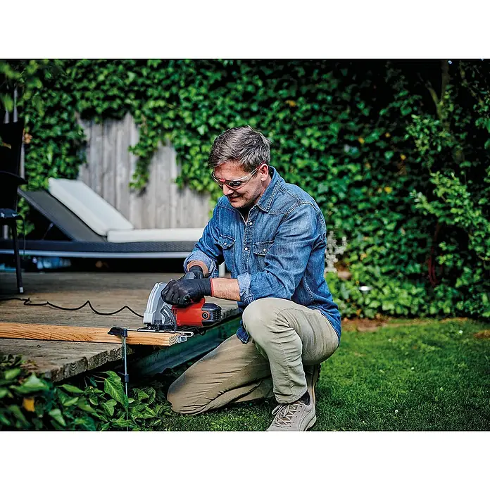 Einhell Handkreissäge TC-CS 1410 - 1.410 W, Durchmesser Sägeblatt: 190 mm, Schnitttiefe bei 90 °: 67 mm - Ein Mann benutzt eine rote und graue Kreissäge im Freien, um ein Holzbrett zu schneiden, während er eine Schutzbrille und Handschuhe trägt.