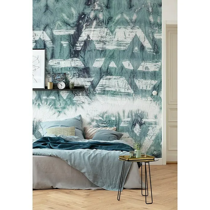Komar Ink Fototapete Tribal Dye - 7 -tlg., B x H: 350 x 280 cm, Vlies - Schlafzimmer mit einem Bett, das mit hellblauer Bettwäsche und Kissen dekoriert ist, vor einer Wand mit einem auffälligen geometrischen Muster.