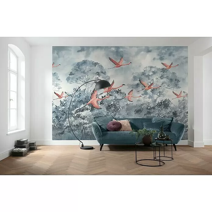 Komar Ink Fototapete Flamingos in the Sky - 8 -tlg., B x H: 400 x 280 cm, Vlies - Wandtapete mit fliegenden rosa Flamingos und grauen Aquarellbäumen, an einer Wand hinter einem blauen Sofa.