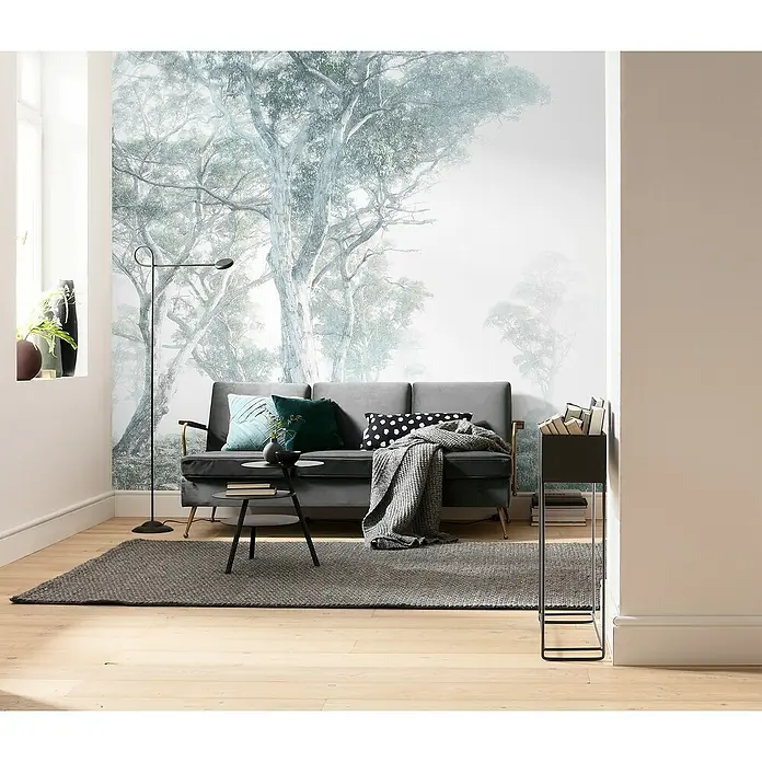 Komar Raw Fototapete Magic Trees - 3 -tlg., B x H: 300 x 280 cm, Vlies - Wohnzimmer mit grauem Sofa, Stehlampe, Teppich und Bücherregal vor einer Tapete mit Baumdesign für eine gemütliche Atmosphäre.