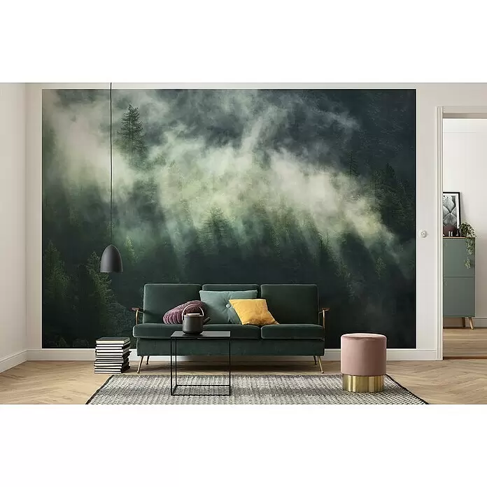 Komar Home Fototapete Misty Crowns - 7 -tlg., B x H: 350 x 250 cm, Vlies - Wohnzimmer mit einem Wandbild eines nebligen Waldes, einem dunkelgrünen Sofa und einem rosa Hocker.