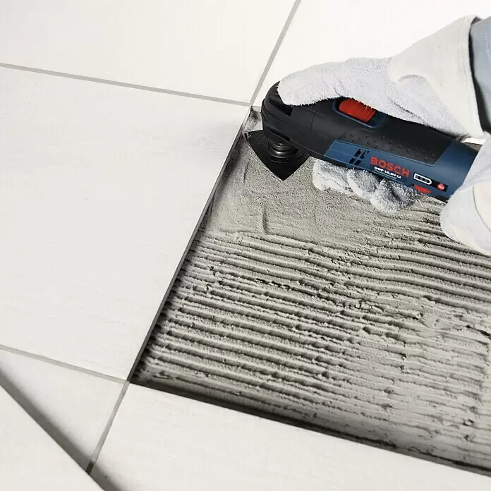 Bosch Zubehör-Set Starlock Best of Cutting - 5 Stk. - Eine behandschuhte Hand verwendet ein blau-schwarzes Bosch Multitool, um Material zwischen weißen Fliesen zu entfernen, wodurch gerillter Kleber sichtbar wird.
