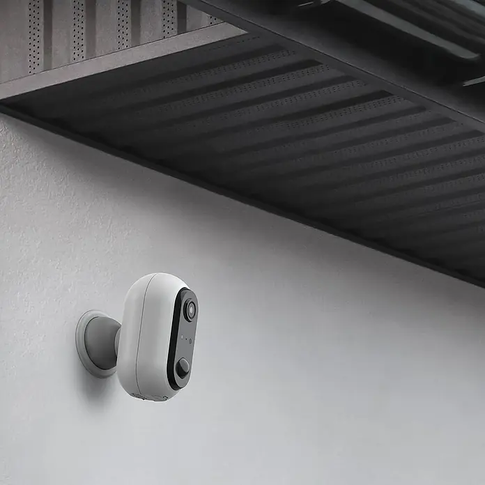 Überwachungskamera HomeCam Akku Outdoor - L x B x H: 55 x 50 x 90 mm - Eine kabellose Überwachungskamera in Weiß ist an einer Außenwand unter einem Vordach befestigt und bietet Sicherheit.