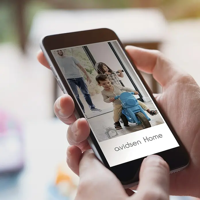 Überwachungskamera HomeCam Akku Outdoor - L x B x H: 55 x 50 x 90 mm - Ein Smartphone zeigt die Avidsen Home App mit einer Live-Aufnahme von spielenden Kindern, die das Smart-Home-Erlebnis hervorhebt.