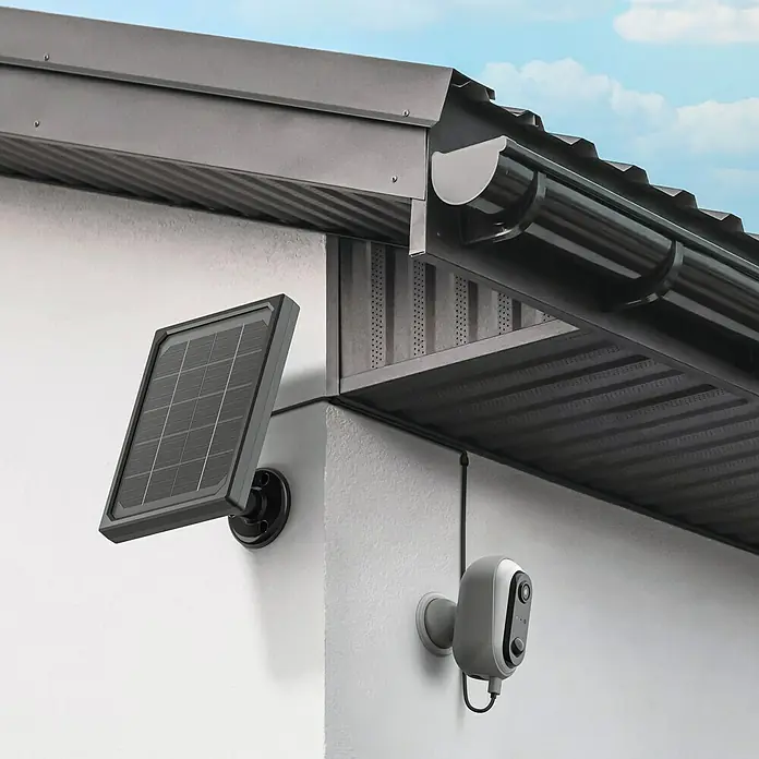 Überwachungskamera HomeCam Akku Outdoor - L x B x H: 55 x 50 x 90 mm - Eine solarbetriebene Überwachungskamera ist an einer weißen Wand unter einem Dachvorsprung montiert.